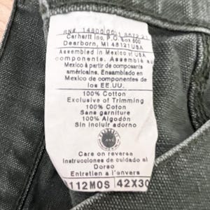 Może przedstawiać: Zbliżenie na zieloną metkę Carhartt z białym tekstem. Metka zawiera nazwę firmy, adres i informacje o materiałach i montażu odzieży. Metka zawiera r&oacute;wnież tekst "100% Bawełna", "Exclusive of Trimming", "Care on reverse", "Instrucciones de cuidado al Dorso", "Entretien a l'onvers", "112MOS" i "42X30".