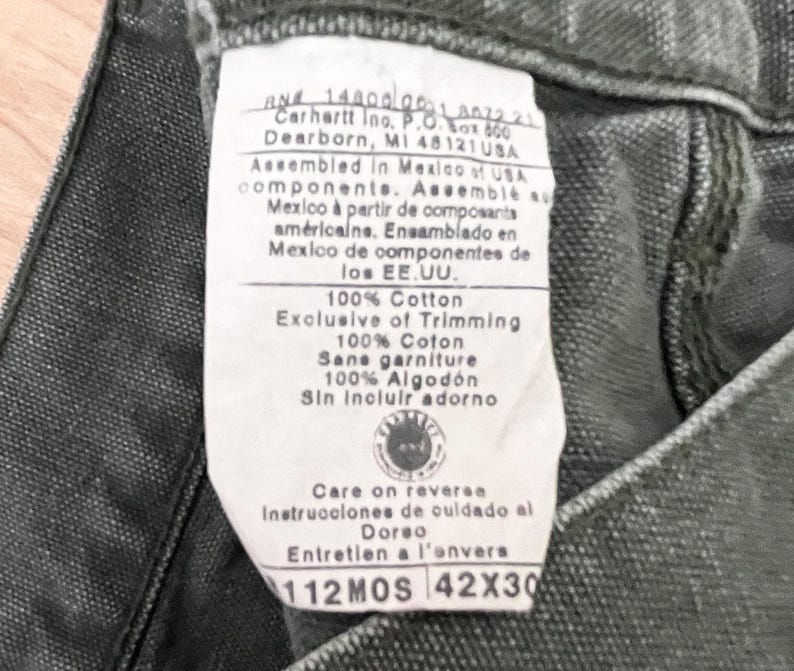 Może przedstawiać: Zbliżenie na zieloną metkę Carhartt z białym tekstem. Metka zawiera nazwę firmy, adres i informacje o materiałach i montażu odzieży. Metka zawiera r&oacute;wnież tekst "100% Bawełna", "Exclusive of Trimming", "Care on reverse", "Instrucciones de cuidado al Dorso", "Entretien a l'onvers", "112MOS" i "42X30".