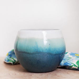 May include: A round ceramic bowl with a gradient of colors. The top is white, transitioning to light blue, then a deeper teal, and finally a dark blue at the base. The bowl sits on a wooden surface.
