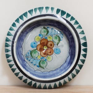 May include: A small, white ceramic bowl with a blue and green rim. The bowl is decorated with a hand-painted orange flower with green leaves and blue accents.