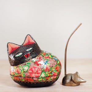 May include: A decorative cat-shaped trinket box with a black head and a body adorned with colorful floral patterns. A small, bronze-colored mouse-shaped object with a curved metal tail stands nearby. The cat box is approximately 4 inches long.