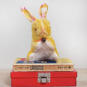 May include: A yellow plush bunny rabbit with pink ears and a metal bell around its neck. The bunny is sitting on top of a red metal box with a book on top of the box. The book is titled "LAROUSSE".