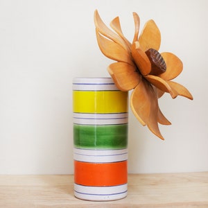 May include: A white ceramic vase with yellow, green, and orange stripes. A wooden flower with a brown center is in the vase.