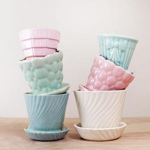 May include: A collection of six ceramic flower pots in various pastel colors, including pink, blue, and white. The pots have unique textures and designs, such as a bow, bubbles, and woven patterns. Each pot sits on a matching saucer.