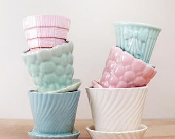 May include: A collection of six ceramic flower pots in various pastel colors, including pink, blue, and white. The pots have unique textures and designs, such as a bow, bubbles, and woven patterns. Each pot sits on a matching saucer.