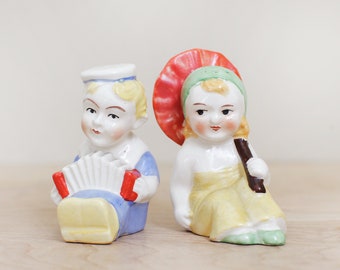 May include: A pair of vintage salt and pepper shakers, one depicting a boy playing an accordion and the other a girl holding an umbrella. The boy is wearing a blue and white sailor outfit, and the girl is wearing a yellow dress with a green hat.