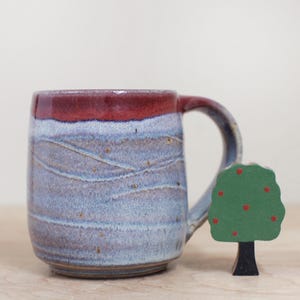 May include: A handmade ceramic mug with a burgundy rim and handle, and a blue and purple body with a wavy design. A small wooden tree with red dots is next to the mug. The mug is on a light brown surface.