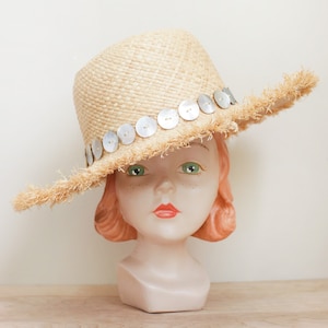 May include: A straw hat with a wide brim and a fringe trim. The hat is decorated with a row of silver buttons.
