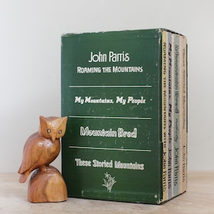 May include: A wooden owl figurine next to a collection of green hardcover books by John Parris. The book titles include "Roaming the Mountains," "My Mountains, My People," and "Mountain Bred."