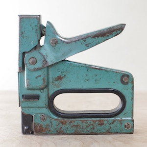 May include: A vintage teal blue staple gun with a black rubber handle and a metal lever. The staple gun has a worn and weathered appearance.