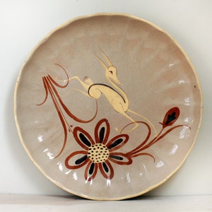 May include: A round, beige ceramic plate with a scalloped edge. The plate features a running gazelle design in cream and brown, with a large flower and floral accents. The plate has a glossy finish.