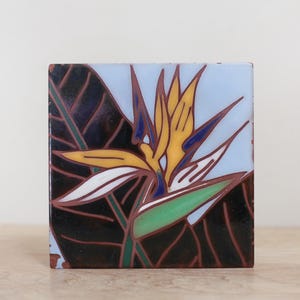 May include: A square ceramic tile featuring a stylized bird of paradise flower illustration. The design includes vibrant yellow, orange, and blue petals against a dark brown leaf background, all outlined in thin, dark brown lines on a light blue field.