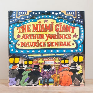 May include: A vintage children's book cover featuring a colorful illustration of a movie theater marquee with the title "The Miami Giant" by Arthur Yorinks and Maurice Sendak. The illustration depicts a group of people in front of the theater.