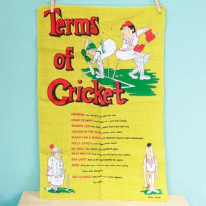 May include: A yellow linen tea towel with the words "Terms of Cricket" in red and black. The towel features cartoon illustrations of cricket players and cricket terms. The towel is hanging from a clothesline.