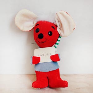 May include: A plush red mouse toy with large cream-colored ears and a white tuft of hair. The mouse wears a gray shirt and red boots, holding a card that reads "CAROLS". A green and white striped scarf is wrapped around its neck.