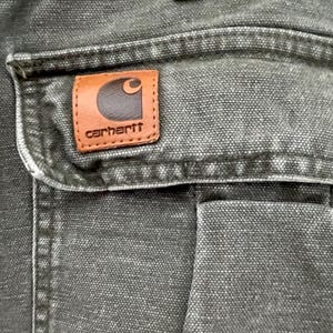 Może przedstawiać: Zbliżenie na ciemnoszarą kieszeń z pł&oacute;tna z brązową sk&oacute;rzaną naszywką Carhartt. Naszywka ma białe logo i napis "carhartt" białymi literami.