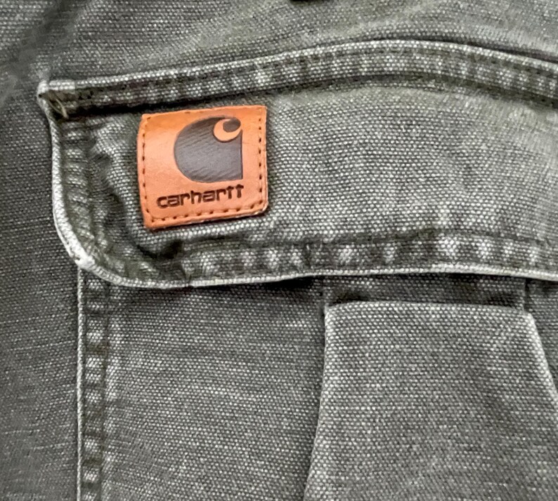 Może przedstawiać: Zbliżenie na ciemnoszarą kieszeń z pł&oacute;tna z brązową sk&oacute;rzaną naszywką Carhartt. Naszywka ma białe logo i napis "carhartt" białymi literami.