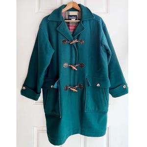 May include: A teal wool coat with a classic toggle closure and large pockets. The coat features a plaid lining and wooden toggles. The coat is hanging on a wooden hanger against a white door.