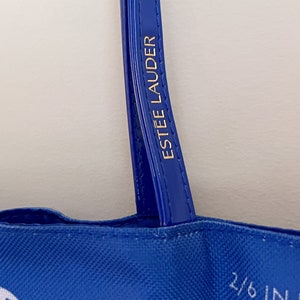 May include: Blue tote bag with white text and a blue handle. The handle has the text "ESTEE LAUDER" in gold lettering.
