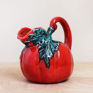May include: A glossy red ceramic pitcher with a rounded body and a curved handle. The pitcher is adorned with green and black leaf accents near the spout. The pitcher is sitting on a wooden surface.