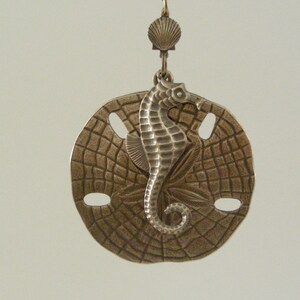 May include: A bronze-toned metal earring featuring a circular sand dollar disc with a textured surface and a central seahorse figure. The earring is suspended from a small, decorative shell-shaped component attached to a metal hook for hanging.