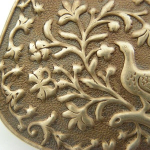 May include: A close-up of a silver-toned metal pendant with an intricate design of a bird perched on a branch with leaves and flowers. The pendant has a textured surface and a slightly aged patina.