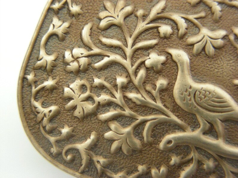 May include: A close-up of a silver-toned metal pendant with an intricate design of a bird perched on a branch with leaves and flowers. The pendant has a textured surface and a slightly aged patina.