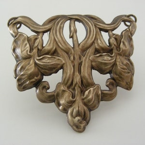 May include: A vintage silver-toned brooch featuring a stylised floral design with three flowers and leaves. The brooch is made of metal and has a detailed, textured surface.