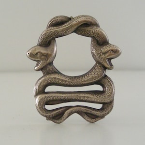 May include: A silver-toned metal belt buckle shaped like two snakes intertwined in a circle. The snakes have detailed scales and open mouths.