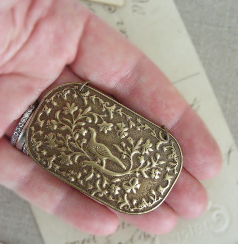 May include: A bronze pendant with an intricate design of a bird perched on a branch with leaves and flowers. The pendant is in the shape of a rectangle with rounded corners.