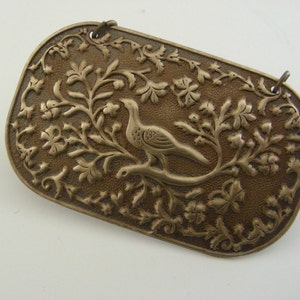 May include: A silver-toned metal plaque with an intricate design of a bird perched on a branch surrounded by leaves and flowers.
