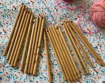 May include: Several wooden crochet hooks with etched patterns, next to two balls of pink yarn. The hooks are displayed on a fabric with a leaf design in blue, orange, and brown.