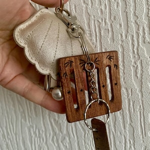 Könnte beinhalten: Ein Schlüsselanhänger mit einem hellgoldenen muschelförmigen Anhänger, einem quadratischen Holzanhänger mit Palmenmotiven und einem kleinen Anhänger. Der Schlüsselanhänger ist an einem silbernen Verschluss und Schlüsselring befestigt.