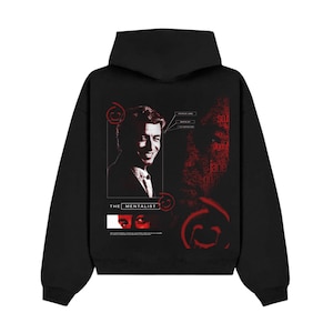 Op de afbeelding: Een zwarte hoodie met een grafische print op de achterkant. Het ontwerp bevat een portret van een man, de tekst THE MENTALIST en rode cirkelvormige symbolen. Aanvullende tekstelementen zijn onder meer PATRICK JANE en MENTALIST.
