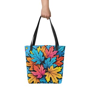 May include: A tote bag with a vibrant design of colorful maple leaves in shades of blue, orange, pink, and yellow against a light blue background. The bag has black handles and is held by a person.