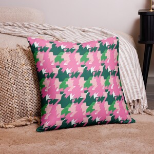 Könnte beinhalten: Quadratisches Kissen mit einem Hahnentrittmuster in Rosa, Grün und Weiß. Das Kissen liegt auf einem beigen Teppich, im Hintergrund sind ein Bett und ein Nachttisch zu sehen. Das Design ist ein sich wiederholendes geometrisches Muster.