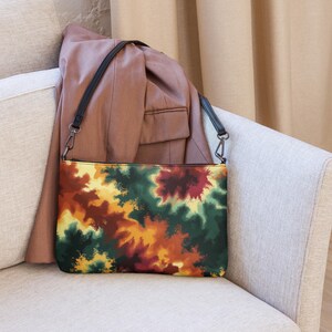 Op de afbeelding: Een kleurrijke clutch met een zwarte schouderriem. De tas heeft een levendig abstract ontwerp in groen, geel, oranje en rood. De tas ligt op een lichtgekleurde bank, met een bruine blazer op de achtergrond.