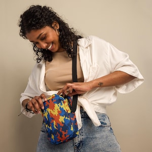 May include: A blue, orange, and yellow pixelated crossbody bag with a black strap. The bag is being opened by a person wearing a white shirt and blue jeans.