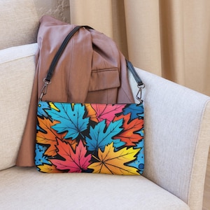 Op de afbeelding: Een rechthoekige clutch met een zwarte band, met een levendig patroon van gestileerde herfstbladeren in blauw, oranje, roze en geel tegen een zwarte achtergrond. De tas ligt op een lichtgekleurde bank, met een bruine jas op de achtergrond.