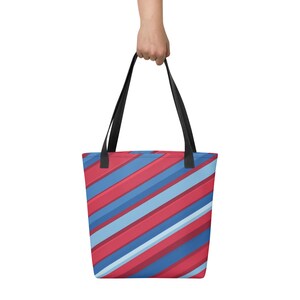 May include: A tote bag with a vibrant diagonal striped pattern in red, blue, and light blue. The bag has black handles and is held up by a hand. This reusable tote bag is suitable for shopping or carrying everyday items.