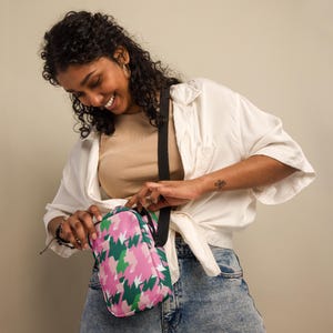 May include: A pink, green, and white patterned crossbody bag with a black strap. The bag is rectangular and has a zipper closure. The bag is being opened by a person wearing a white shirt and blue jeans.