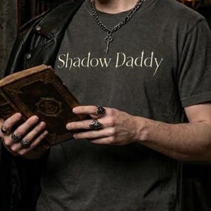 May include: A dark gray t-shirt with the text "Shadow Daddy" in white script. The person is wearing a black leather jacket and black jeans. They are holding an old book with a detailed cover and wearing rings.