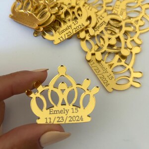 May include: A collection of gold-colored acrylic crown-shaped charms with a reflective finish. Each charm features the engraved text Emely 15 and the date 11/23/2024. The crown design has a central point and rounded decorative cutouts throughout.