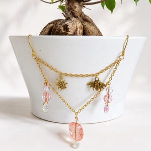 May include: A gold-toned metal chain garland featuring two tiers. The upper tier displays a small starburst charm and a bee charm. The lower tier is accented with dangling pink faceted crystal beads and a larger central pink oval crystal bead.
