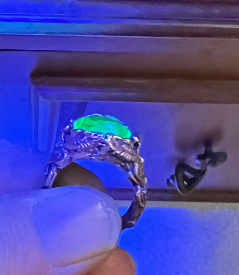 May include: A silver ring with a green gemstone set in a detailed, intricate design. The ring is worn on a finger.