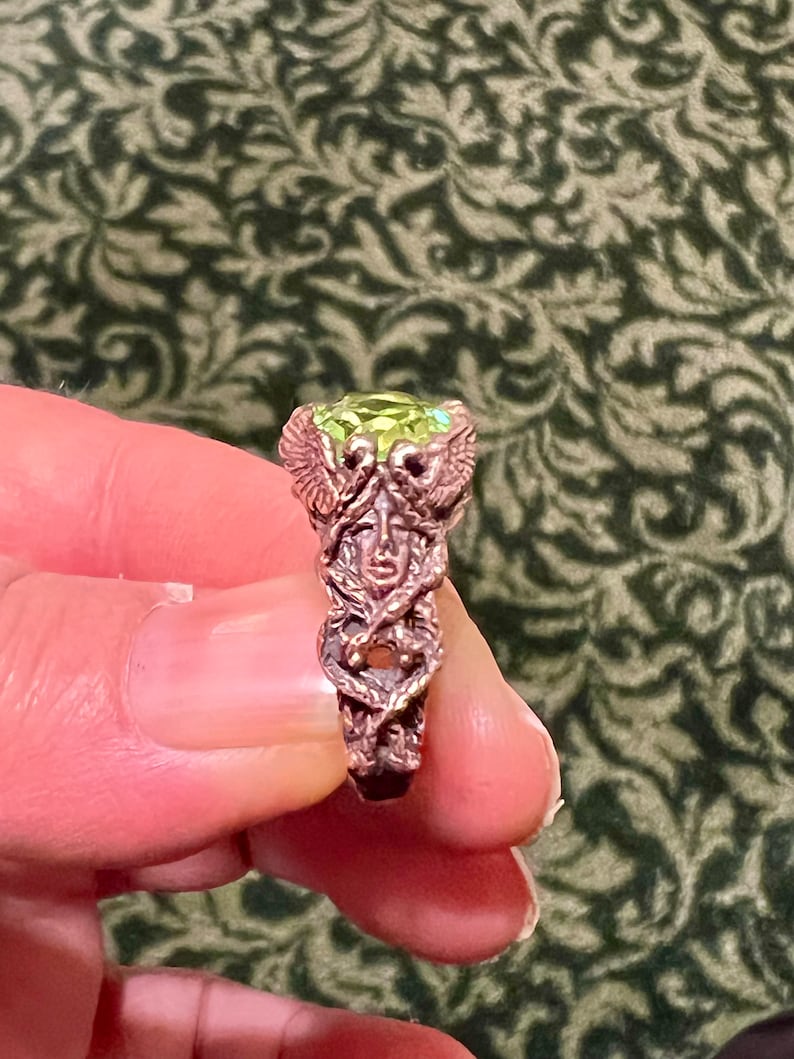 May include: A copper ring with a green gemstone and a detailed design of a winged creature with a human face.