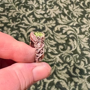 May include: A ring with a green gemstone set in a detailed, intricate design. The ring is made of metal with a rose gold finish.