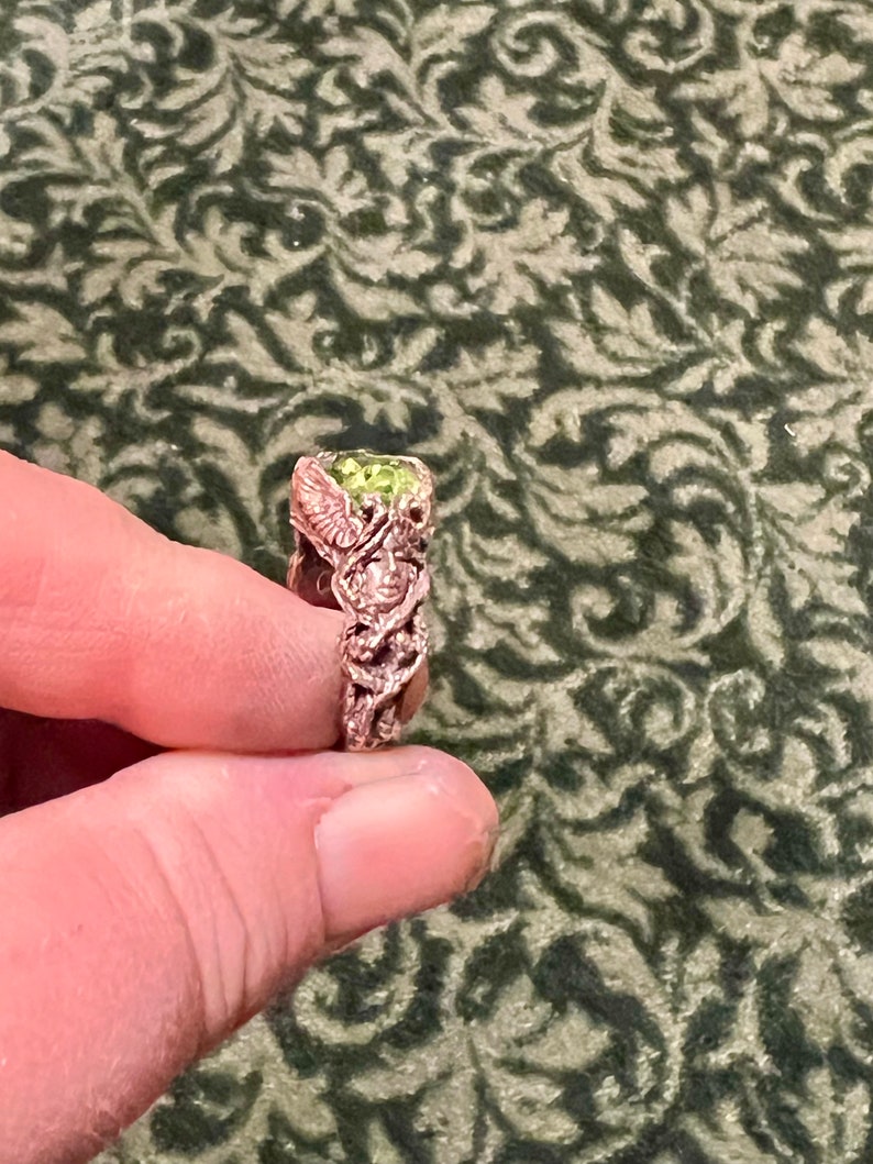 May include: A ring with a green gemstone set in a detailed, intricate design. The ring is made of metal with a rose gold finish.