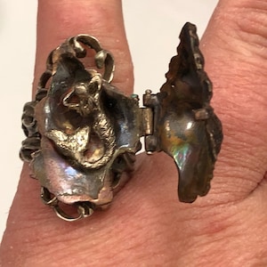 May include: A silver ring with a hinged design. The ring features a detailed, sculpted design of a mermaid with a tail and a crown. The ring is open and the inside is visible.