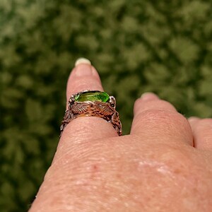 May include: A silver ring with a green gemstone set in a detailed, intricate design. The ring is worn on a finger.